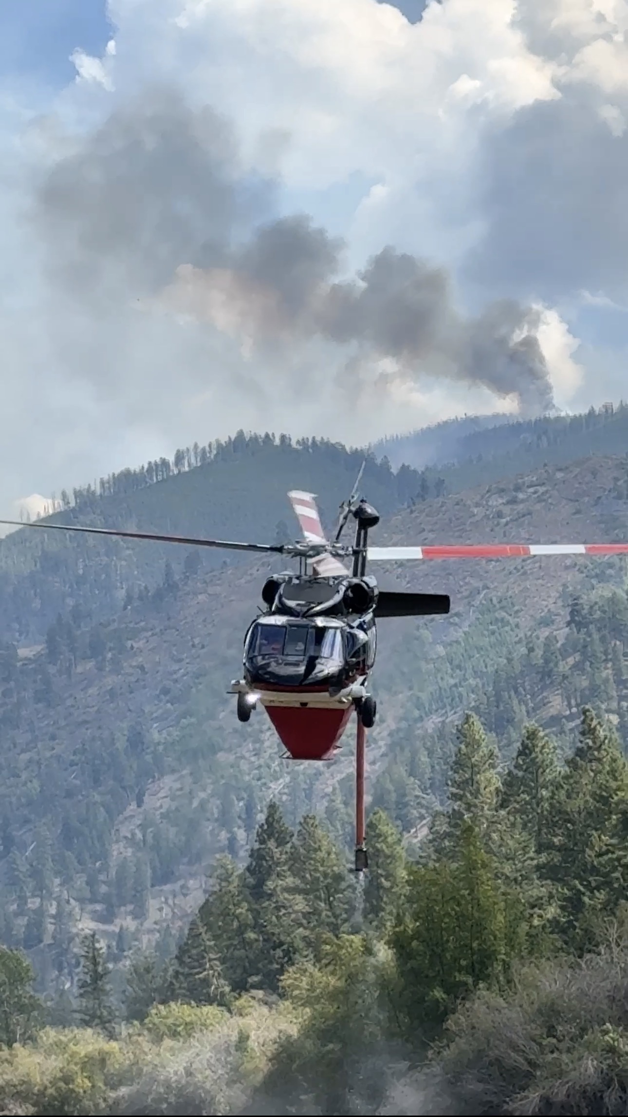 A UH-60 with a belly tank and snorkel hovering near a wildfire